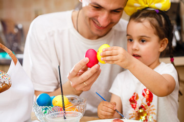 Father and his daughter are painting eggs. Happy family preparing for Easter. Cute little child girl wearing bunny ears.