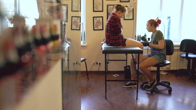 Beautiful red haired woman tattoo artist tattooing picture on leg of young girl client in studio