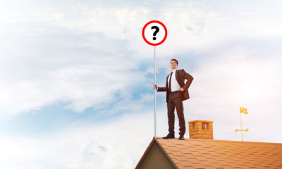 Businessman on brick house roof showing banner with question mark. Mixed media