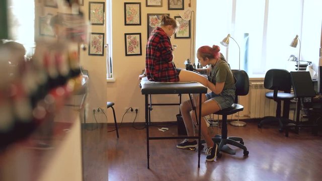 Young female red haired tattoo artist tattooing picture on leg of client over sketch in studio indoors