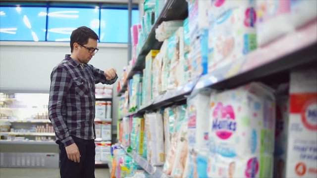 A serious man in glasses, dressed in a plaid shirt examines diapers in the children's department of the hypermarket. A person considers children's belongings for his little child.