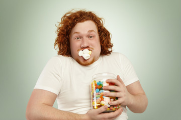Never enough. Close up shot of funny young plump man with curly ginger hair holding tight jar of...