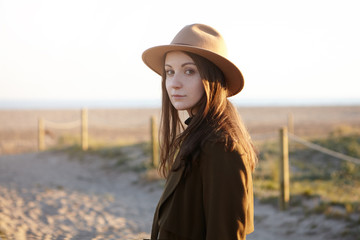 Outdoor portrait of stylish young European woman wearing trendy hat and black coat looking at camera with subtle smile while having nice evening walk by the sea, dreaming and admiring sunset