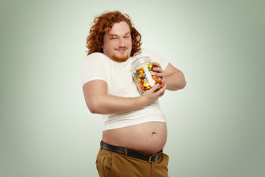 Funny obese young red-haired man with big belly hanging out his undone jeans having happy and cheerful look,holding tight large glass jar of sweets, clasping it to heart. Diet, junk food and calories