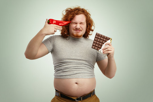 Red-haired young European bearded man wearing shrunk t-shirt with his stomach sticking out of jeans, holding bar of chocolate in one hand and red pepper at his temple, fed up with vegetable diet