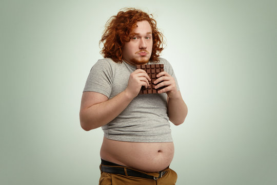 Overweight fat man with ginger curly hair looking indecisive and hesitant, holding large bar of chocolate with both hands while forbidden to eat sugar, and junk food because of strict low-carb diet