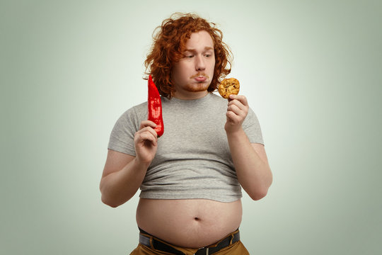People, food, diet, nutrition and unhealthy lifestyle. Studio shot of sad indecisive young plump man with big stomach hanging out of jeans, facing difficult dilemma between vegetable and cookie