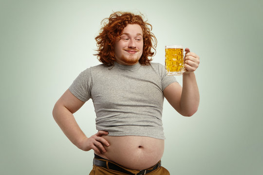Happy redhead overweight man with big belly sticking out of his shrunk t-shirt holding glass of cold beer, looking in anticipation, impatient to feel its good taste while relaxing at home after work