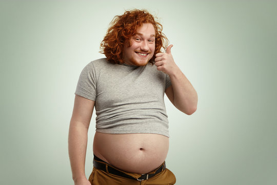 People, nutrition, body shape and healthy lifestyle concept. Happy young plump chubby man with curly ginger hair and beard showing thumps up, saying he is doing okay while his belly sticking out