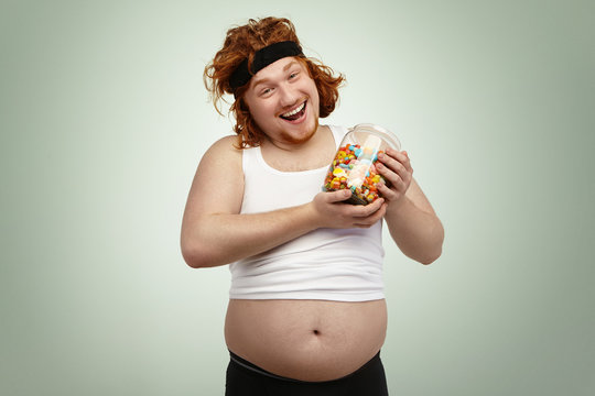 Happy handsome red-haired man wearing hairband and shrunk tank top holding glass jar of candies, rejoicing at delicious but unhealthy junk foot after cardio training, fighting excess weight