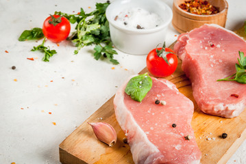 Raw pork, steak, cutlet. Meat, a source of protein. On a cutting board, on a white stone table. With spices, herbs and tomatoes for cooking. Copy space