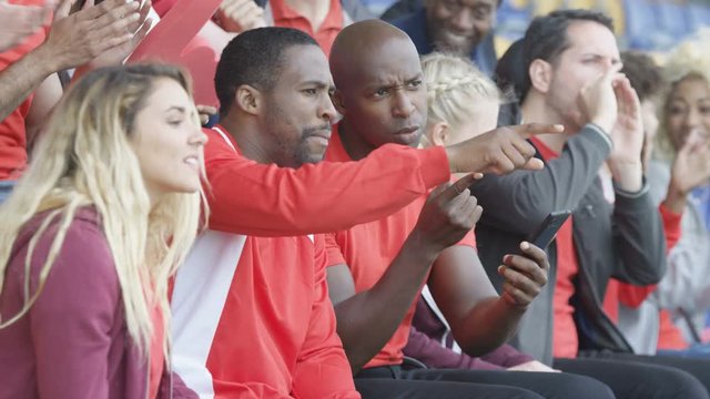  Spectators watching sports game in stadium, 2 men looking at smartphone