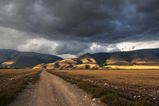 Road through a dry desert steppe to village on a sunset highland mountain plateau with yellow grass with ranges on a horizon, Kurai, Altai, Siberia, Russia