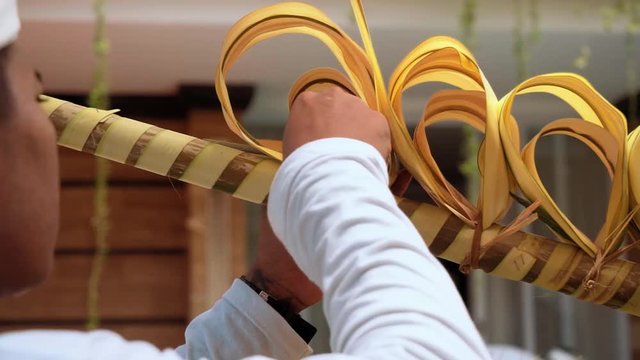 Balinese man decorating a penjor with coconut palm leaf ribbons on Galungan Day (Bali holiday ceremony). Close up shot with Sony a7s on tripod on sunny day