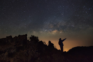 A Man is standing next to the milky way galaxy pointing on a bright star at Doi Luang Chiang Dao with Thai Language top point signs. Long exposure photograph.With grain