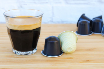 Cup of black coffee with foam and coffee capsules on wooden background.