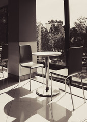 A table and two chairs by bright lit window casting shadows on the ground. Cafe background. Brown tone.