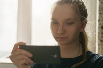 Fototapeta premium teen girl sitting near window with smartphone, toned photo with shallow focus