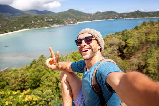 Young male hipster traveler doing selfie overlooking the tropical sea. Adventure, vacation, wonderlust, internet, technology concept.