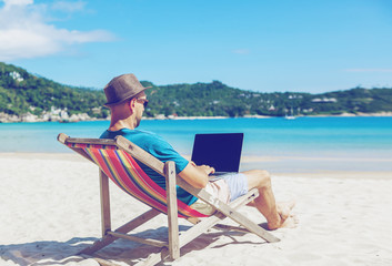 Obraz na płótnie Canvas Young hipster man with laptop on tropical beach. Travel, vacation, internet, freelance job concept