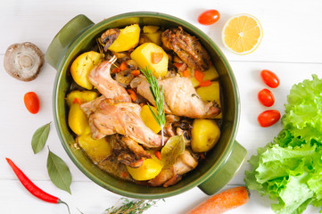 Top flat view of roasted rabbit meat with vegetables in round ceramic pot on white wooden table surface. Food ingridients
