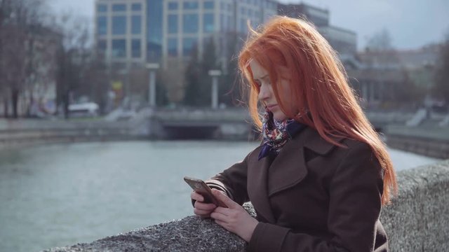 Young redhead woman is standing near the river bank in the city with a cell phone in the hands, looking at the screen, windy day 