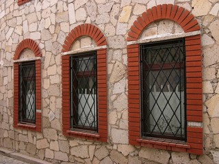 Vergitterte Fenster mit gemauerten Rundbögen aus rotem Backstein in einer Fassade aus Naturstein...
