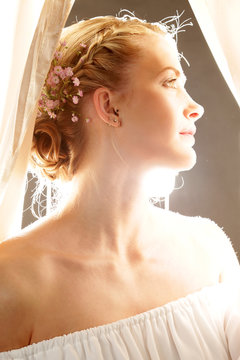Eine Frau mit Blumen im Haar und Zopffrisur in strahlendem Licht zischen Vorhängen stehend