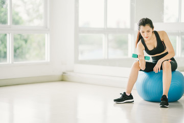 Sexy Asian girl exercising with dumbbell on fitness ball at fitness gym with copy space, sport and healthy lifestyle concept