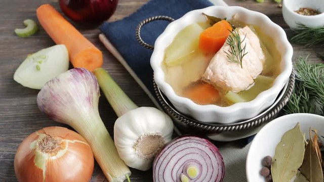 Hot steaming fish soup on wooden table. Roots, vegetables, spices, condiments nearby. Still footage - camera locked down. Closeup.