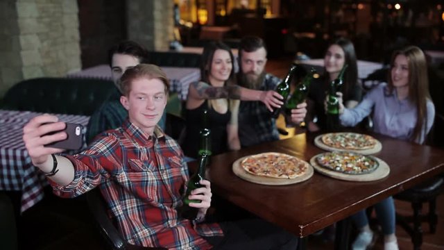 A company of friends resting in a pizzeria and making selfie on the background of pizza and beer
