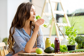 Beautiful woman sitting with drinks and healthy green food at home. Vegan meal and detox concept - obrazy, fototapety, plakaty