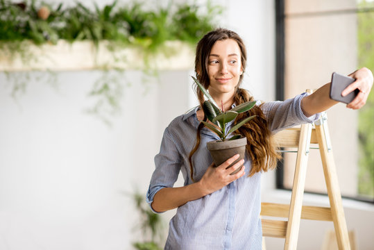 Woman making selfie photo standing with flowerpot on the ladder at home