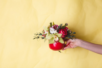 Hand holds a small red vase with flowers and lily on a yellow background