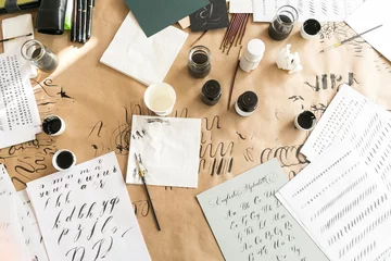 Kalligraphieblätter, Federn, Papier, Tinte und Stifte auf Kraftpapierhintergrund. Kalligraphie-Workshop für Schriftzüge. Buchstaben des englischen Alphabets mit einem Pinsel geschrieben. Konzept-Hobby. Ansicht von oben, flach liegend. © melnikofd