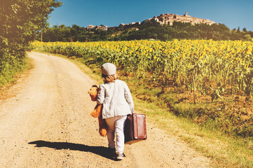 Outdoor portrait of a funny little boy walking down the road in countryside, holding old small suitcase and teddy bear toy, back view. Image taken in Tuscany, Italy