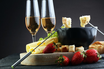 Gourmet Swiss fondue dinner with assorted cheeses on a board and strawberries alongside a heated pot of cheese fondue with two forks dipping bread and white wine behind in a tavern or restaurant