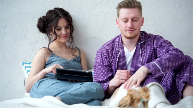 Happy newlyweds who are dressed in homemade clothes and their dog enjoy watching a movie on a tablet. Cute woman with black hair and a young man spend a day off in bed.