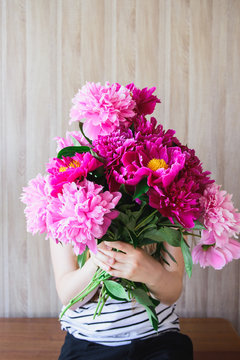 happy white caucasian young woman holding red and pink peonies bouquet in front of face at wood wall backdrop