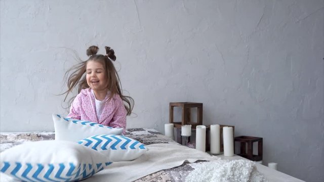 A sweet and joyful girl with funny tails on her head plays with pillows on the bed in the bedroom. A beautiful child jumps and laughs while amusing himself.