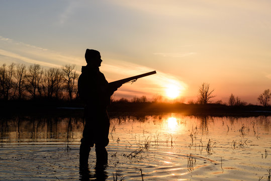 Silhouette of a hunter at sunset in the water with a gun.
