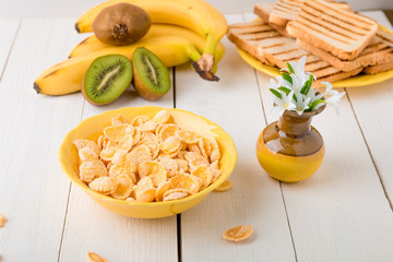 Healthy breakfast with cereal flakes and fruit near vase with flowers on white background. Yellow tone.