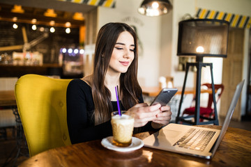 Young woman sitting in coffee shop at wooden table, drinking coffee and using smartphone. On table is laptop. Girl browsing internet, chatting, blogging.