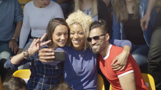  Friends sitting in the crowd at sports event pose to take a selfie with phone