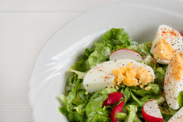 Close-up side view a colorful plate of green salad with lettuce, tomatoes, cucumber, onion, radishes and olives on white wooden board. Healthy food and eating concept. Fresh green salad background.