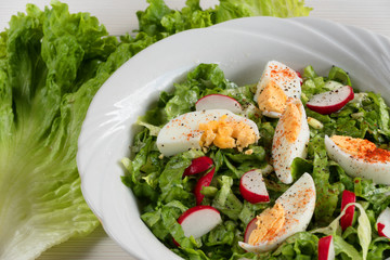 Close-up side view a colorful plate of green salad with lettuce, tomatoes, cucumber, onion, radishes and olives on white wooden board. Healthy food and eating concept. Fresh green salad background.