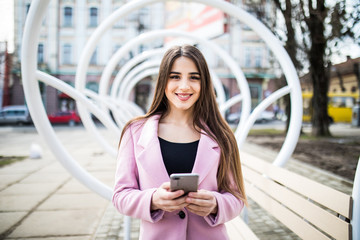 City lifestyle stylish girl using a phone texting on smartphone app in a street near the modern bench