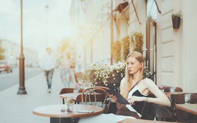 Charming caucasian woman is thoughtfully looking aside while sitting in street cafe with digital tablet, attractive pensive blonde adult lady with touch pad is planing her day during morning breakfast