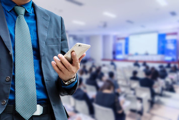 businessman using the mobile phone, blurred of conference hall or seminar room with attendee background.