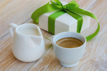 Cup of coffee and small milk jug on rustic wood table, white gift box with silk green ribbon and bow on the background. Good morning surprise concept.